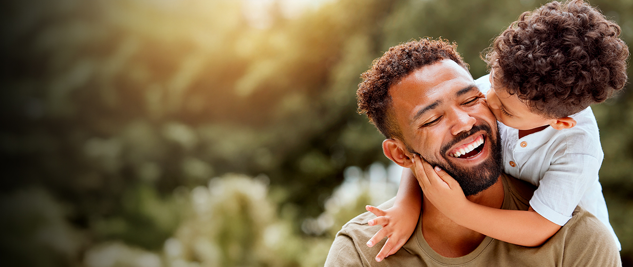 A man with a beard smiles joyfully as a young child with curly hair hugs him and kisses his cheek. They are outdoors with blurred greenery in the background. The mood is warm and affectionate.