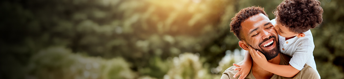 A man with a beard smiles joyfully as a young child with curly hair hugs him and kisses his cheek. They are outdoors with blurred greenery in the background. The mood is warm and affectionate.